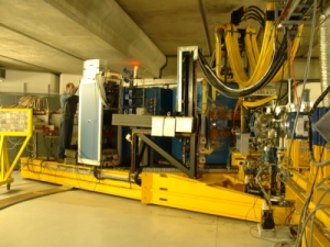 In the picture the K600 magnetic spectrometer is shown. On the right-hand side the beam line, the target chamber and
several vacuum pumbs can be seen. The huge spectrometer consisting of dipole and quadrupole magnets
is placed in the middle of the picture. The focal plane detector system is shown on the left-hand side. It is shielded
with a massive lead against radiation background coming from the beam dump and the scattering chamber. In the picture
is working at the electronics. The spectrometer is painted in blue and in yellow colors.